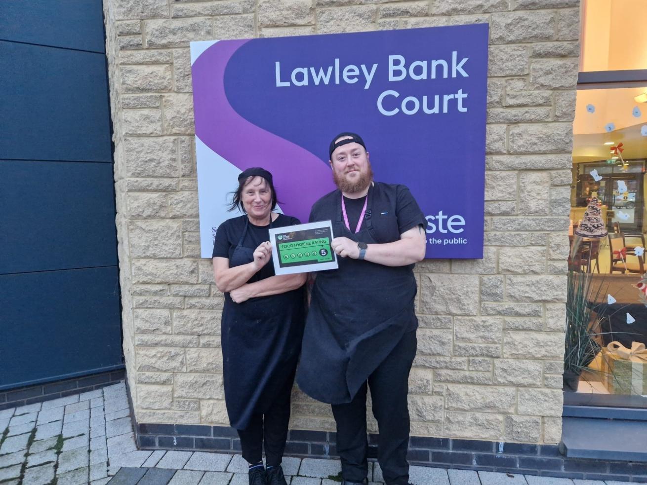 Two members of a kitchen team standing outside Lawley Bank Court, as shown by a purple sign on the building, holding a certificate displaying a '5' food hygiene rating. They are wearing black uniforms and smiling.
