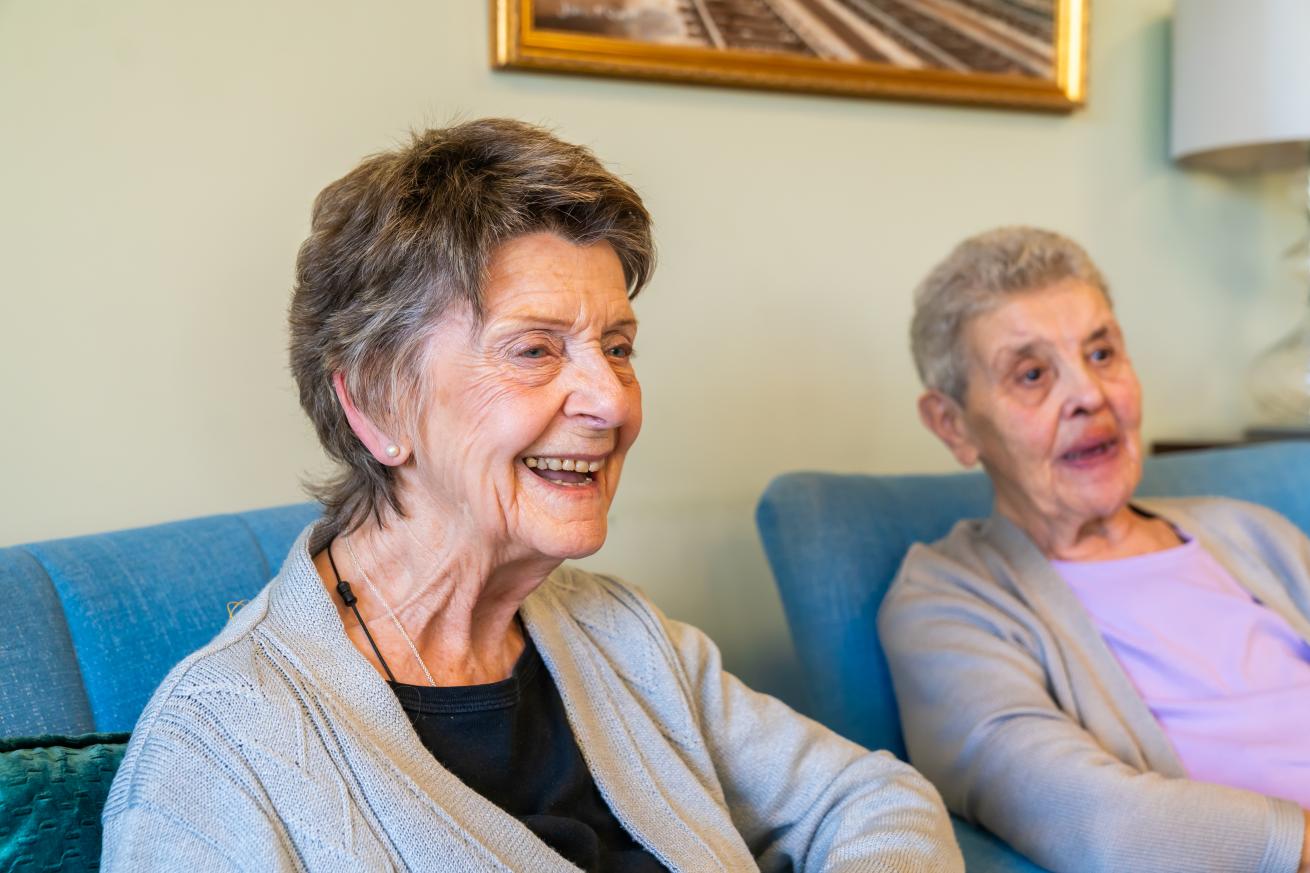 Two elderly women sitting on blue armchairs looking off camera and smiling