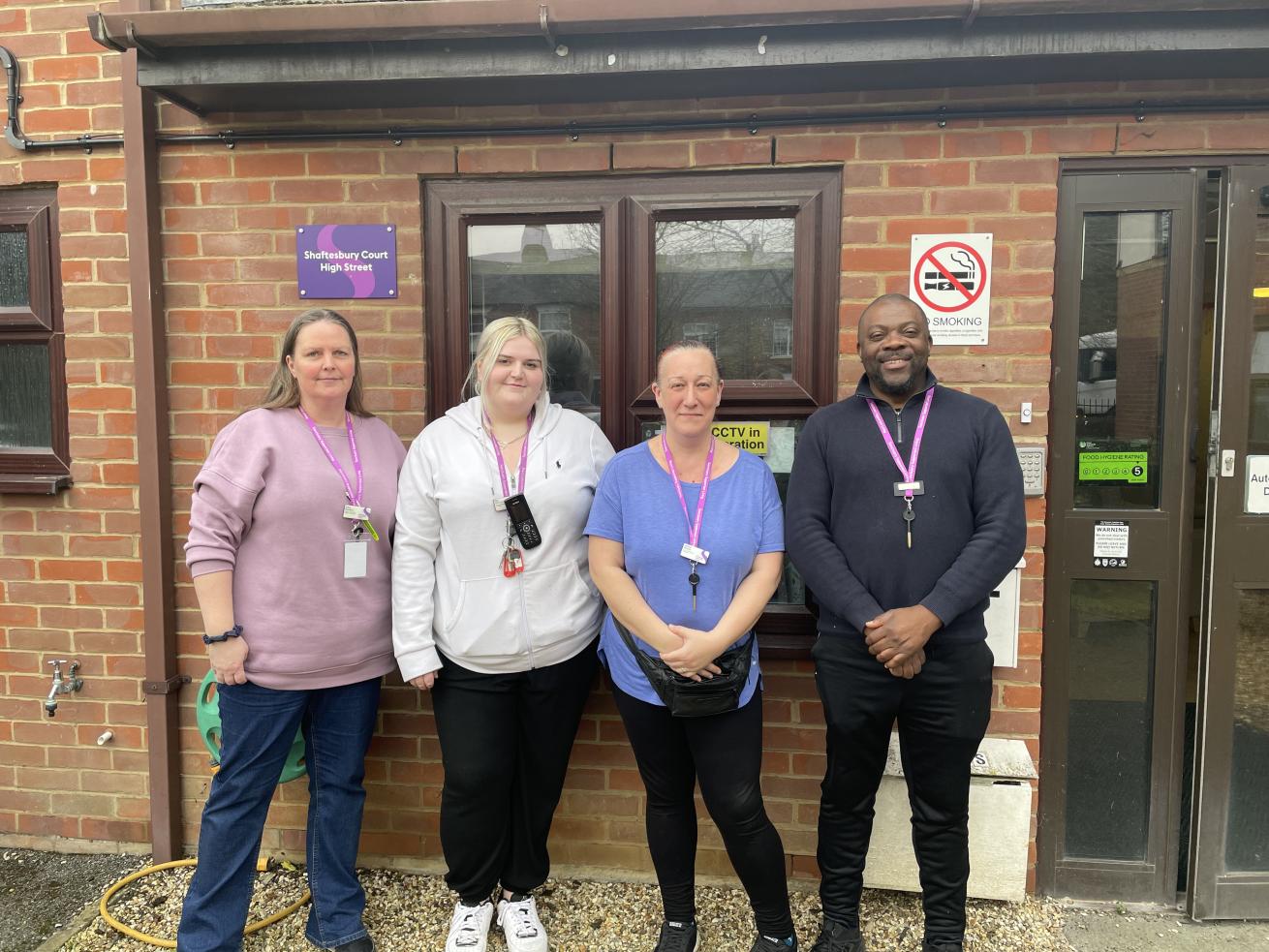 Three white women and one black man standing in a row outside by the front door of a residential care home. They are all wearing purple lanyards and are smiling at the camera. There is a green sticker in the window behind them, showing a rating of five from the Food Standards Agency.