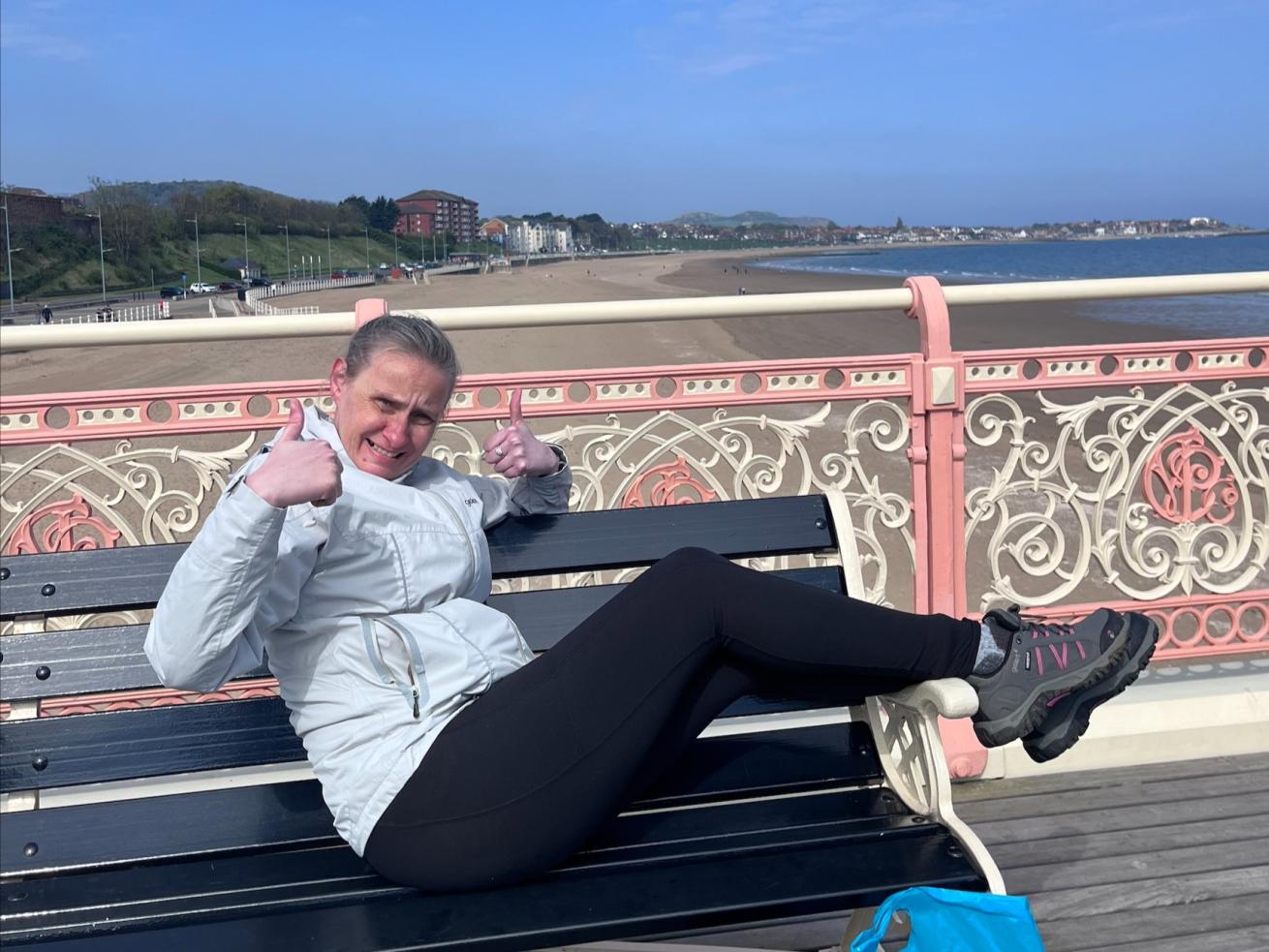 A woman wearing a pale grey jacket, black leggings and trainings, sits along a bench on a pier by a beach, smiling and giving a thumbs up to the camera.