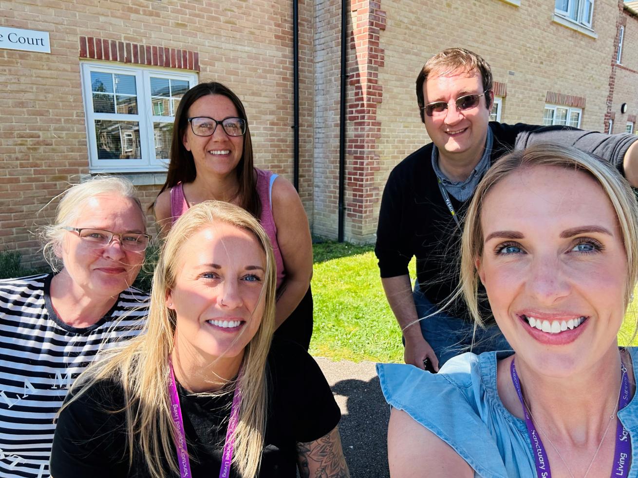 Four women and one man, wearing purple Sanctuary Supported Living lanyards are standing on a lawn outside a large brick building, posing together and smiling for the camera.