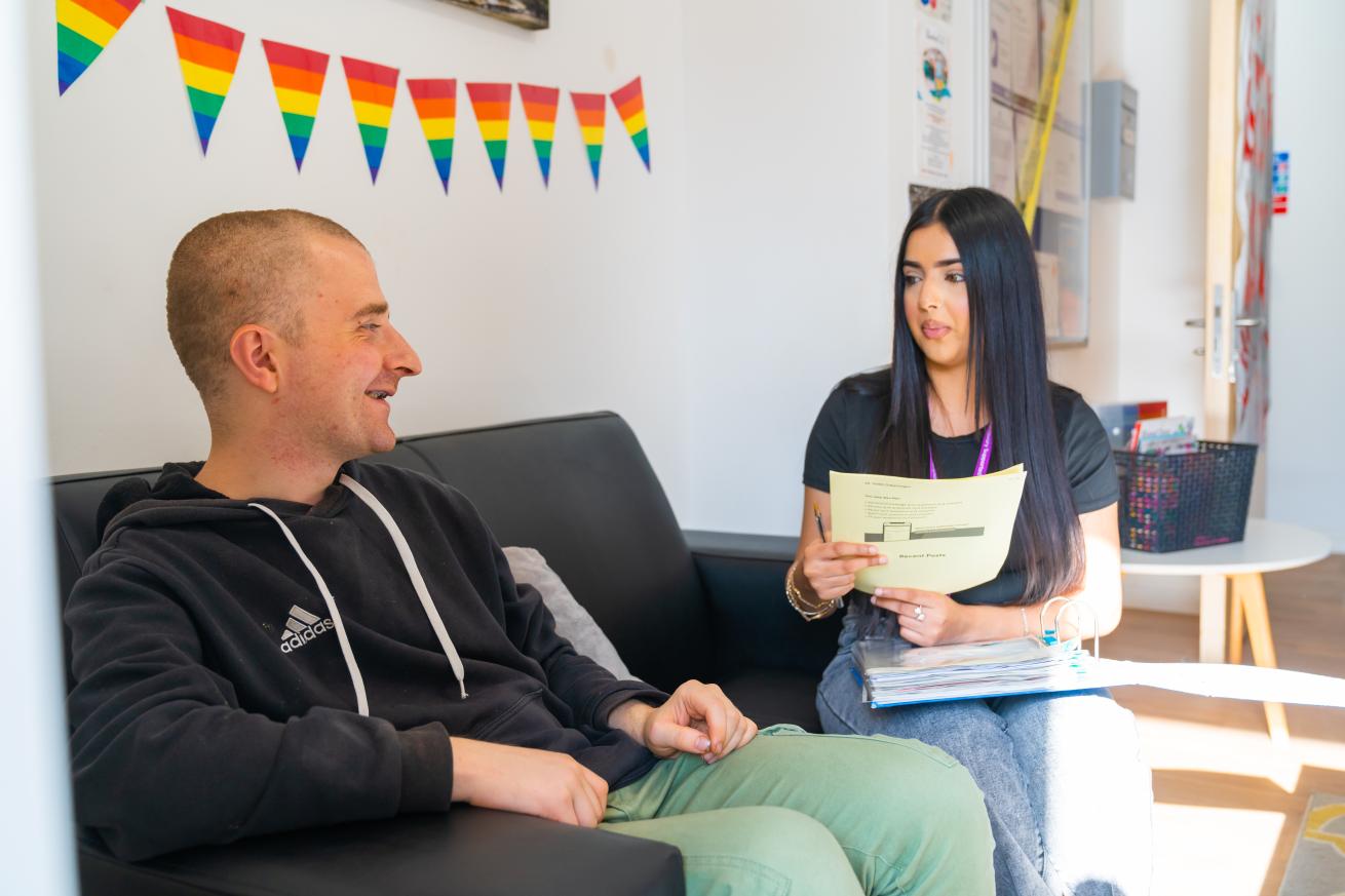 Two young people sat on a sofa one has a binder and is holding some papers