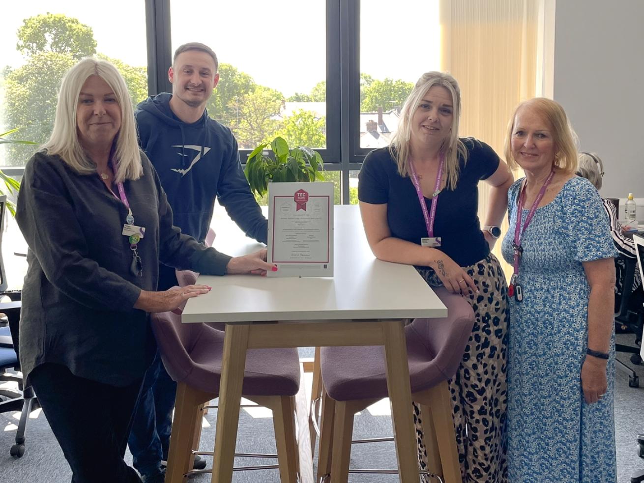 Three women and one man stood around an office table, smiling at the camera and showing off a certificate in the middle of the group