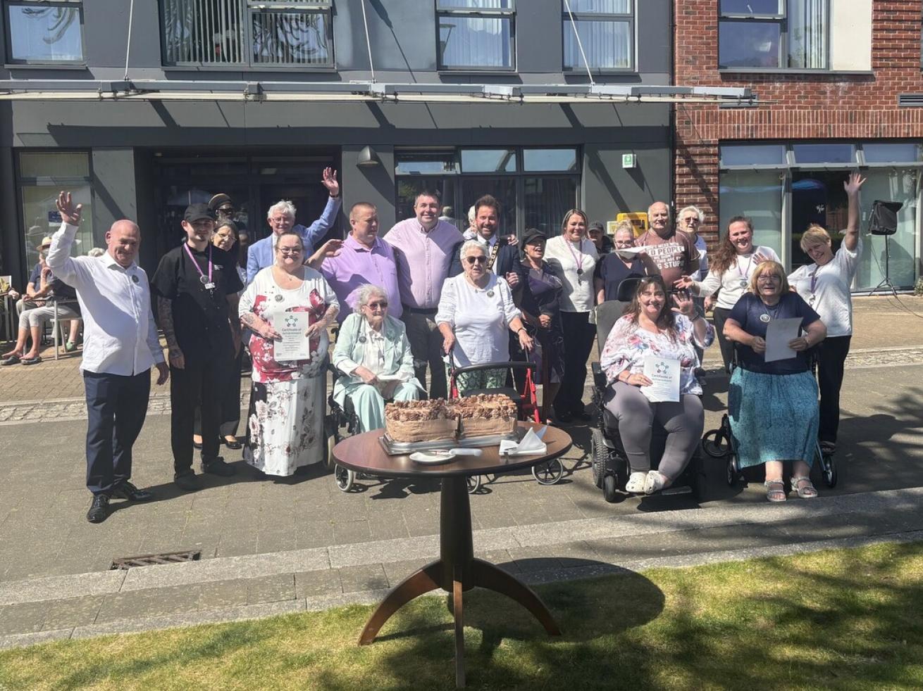 A large group of people stood or sat in wheelchairs all together, posing for the camera. They're in front of the main entrance to a large building, with a table in front of them which has a large rectangular chocolate cake on it. Some people are holding certificates.