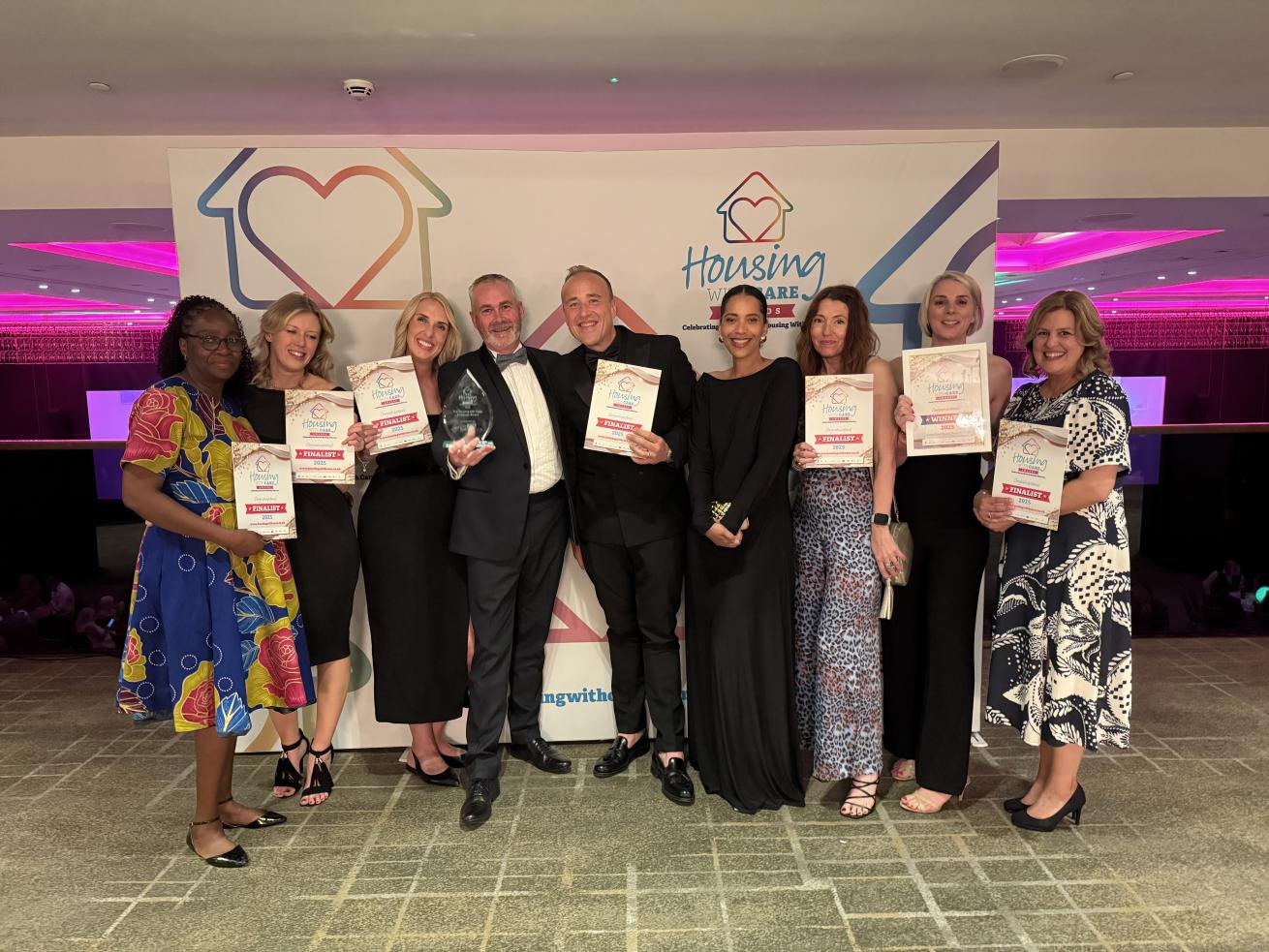 A group of nine people in suits and evening dresses, stood in a row smiling at the camera in a large room, in front of a 'Housing with Care Awards sign'. They are happily showing off their certificates and one glass trophy award.