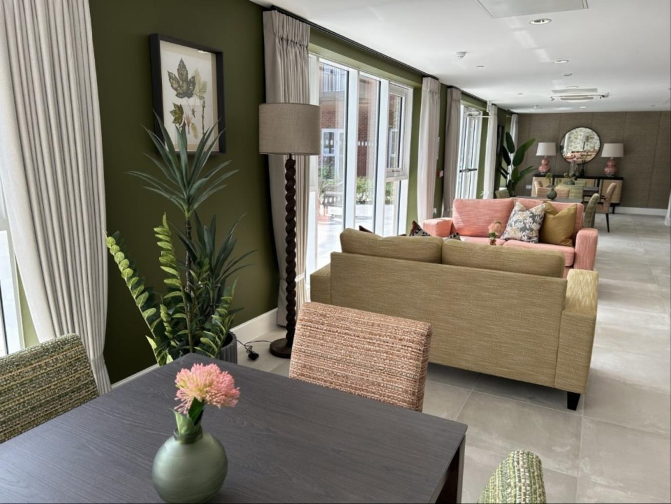 A modern living room with green walls, large windows with white curtains, and a mix of furniture including a beige sofa, pink armchairs, and a dining table. The room is decorated with plants, framed artwork, and various decorative items.