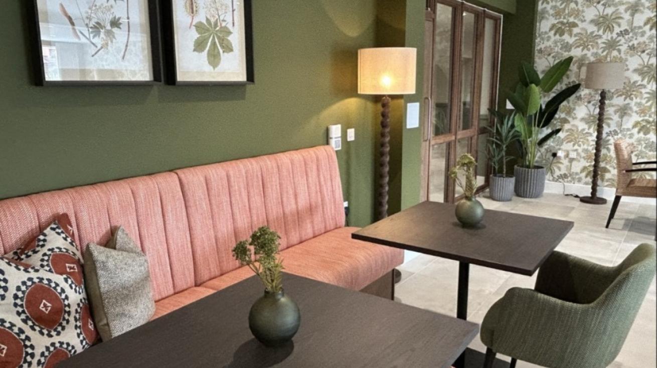 A cozy seating area with a green wall, two framed botanical prints, a pink upholstered bench with decorative pillows, and a green armchair. Two dark wooden tables with small vases of greenery sit in front, alongside a beige-shaded floor lamp. Leaf-patterned wallpaper and glass doors leading to another room complete the background, with several potted plants nearby.