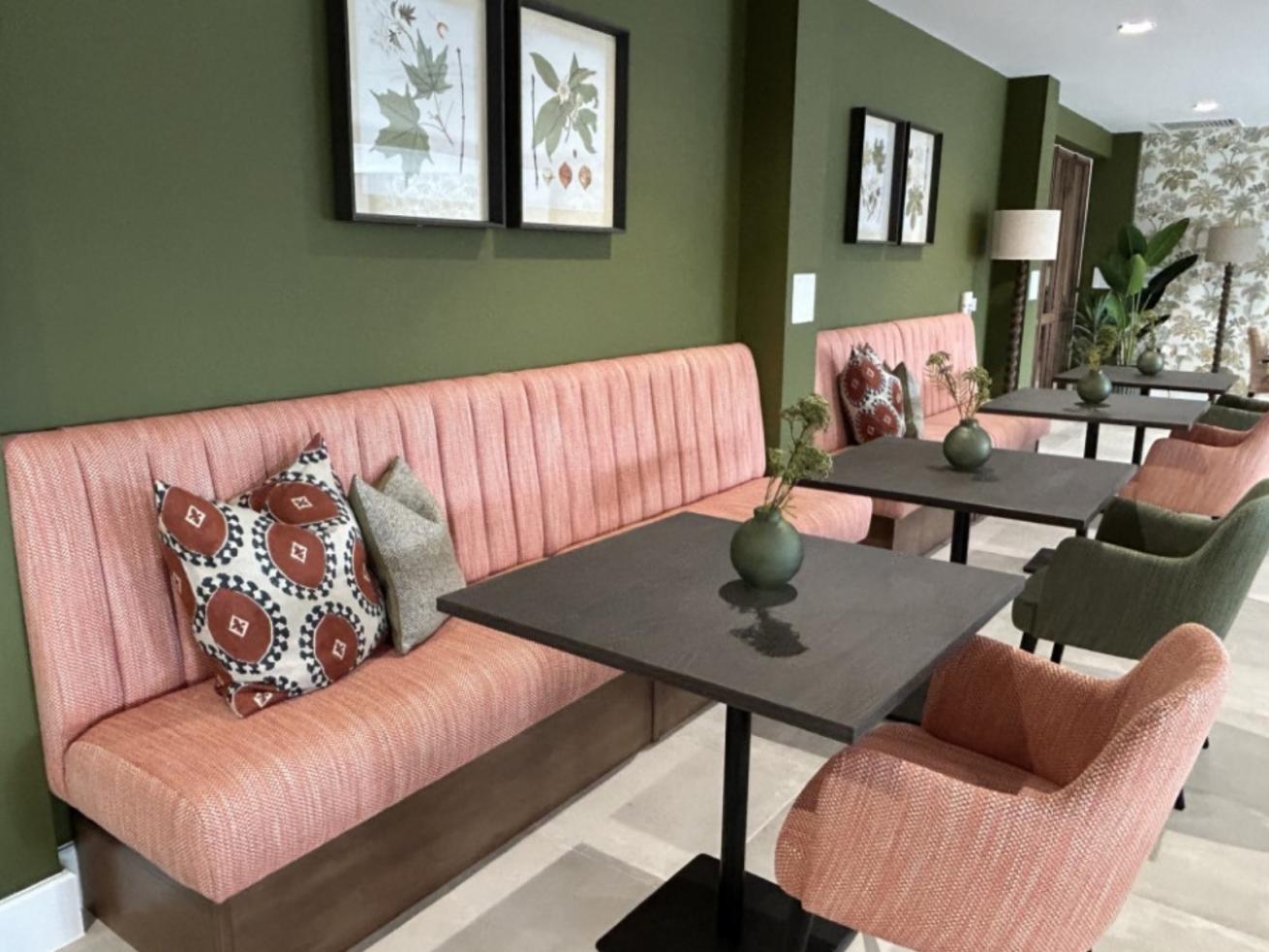 A cozy seating area with pink cushioned benches, green walls, and small tables adorned with green vases containing plants. The space is decorated with framed botanical prints and has a mix of pink and green upholstered chairs.