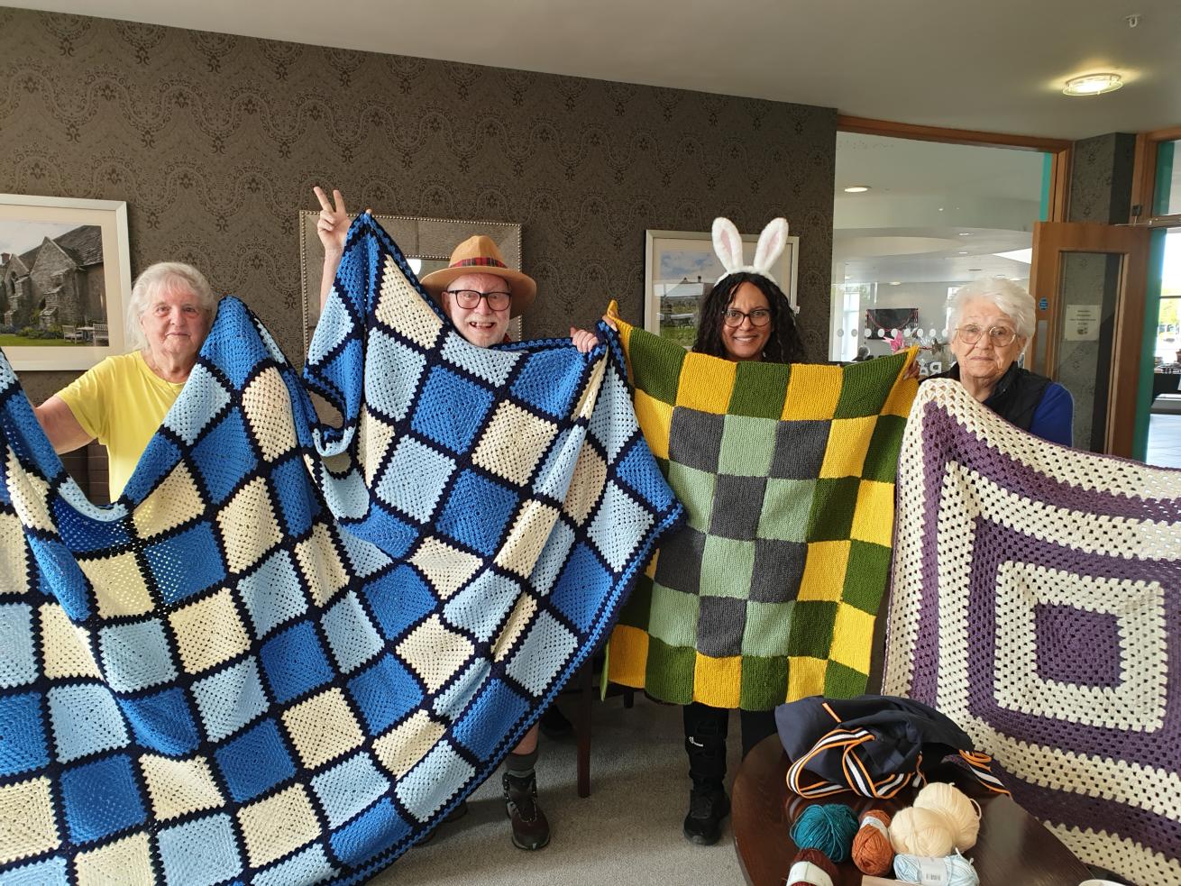 Four older people stood in a communal lounge of a retirement community, smiling at the camera and proudly displaying three knitted and crocheted blankets - one blue and cream, one yellow and green, and one purple and cream.