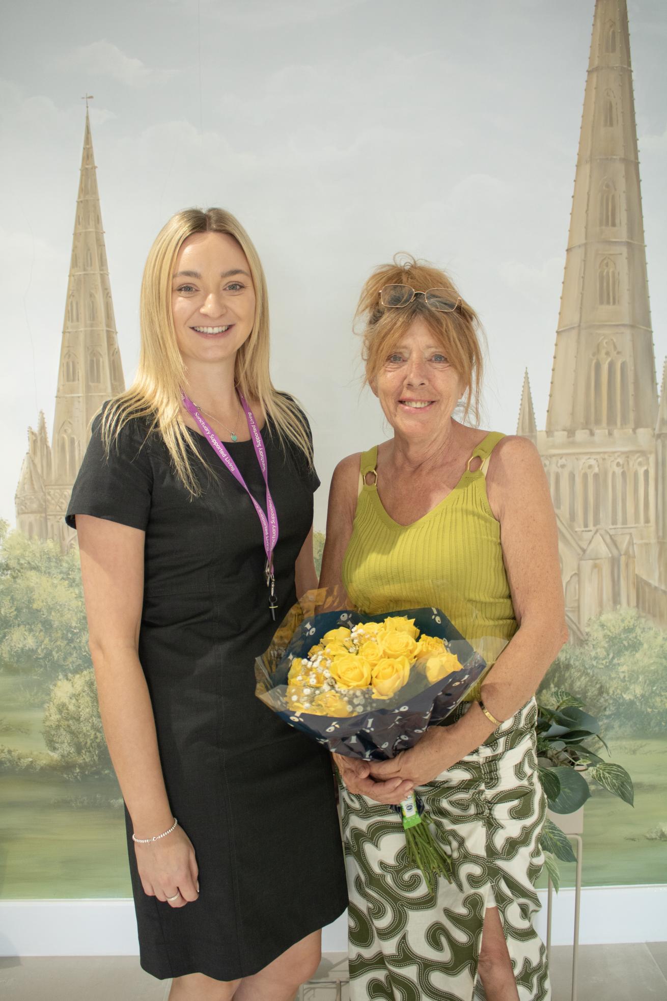 A young woman in a black dress with a lanyard stands next to an older woman wearing a green top and patterned skirt. The older woman is holding a bouquet of yellow roses. They are posing in front of a backdrop featuring tall, pointed church spires and a green landscape. Both women are smiling.
