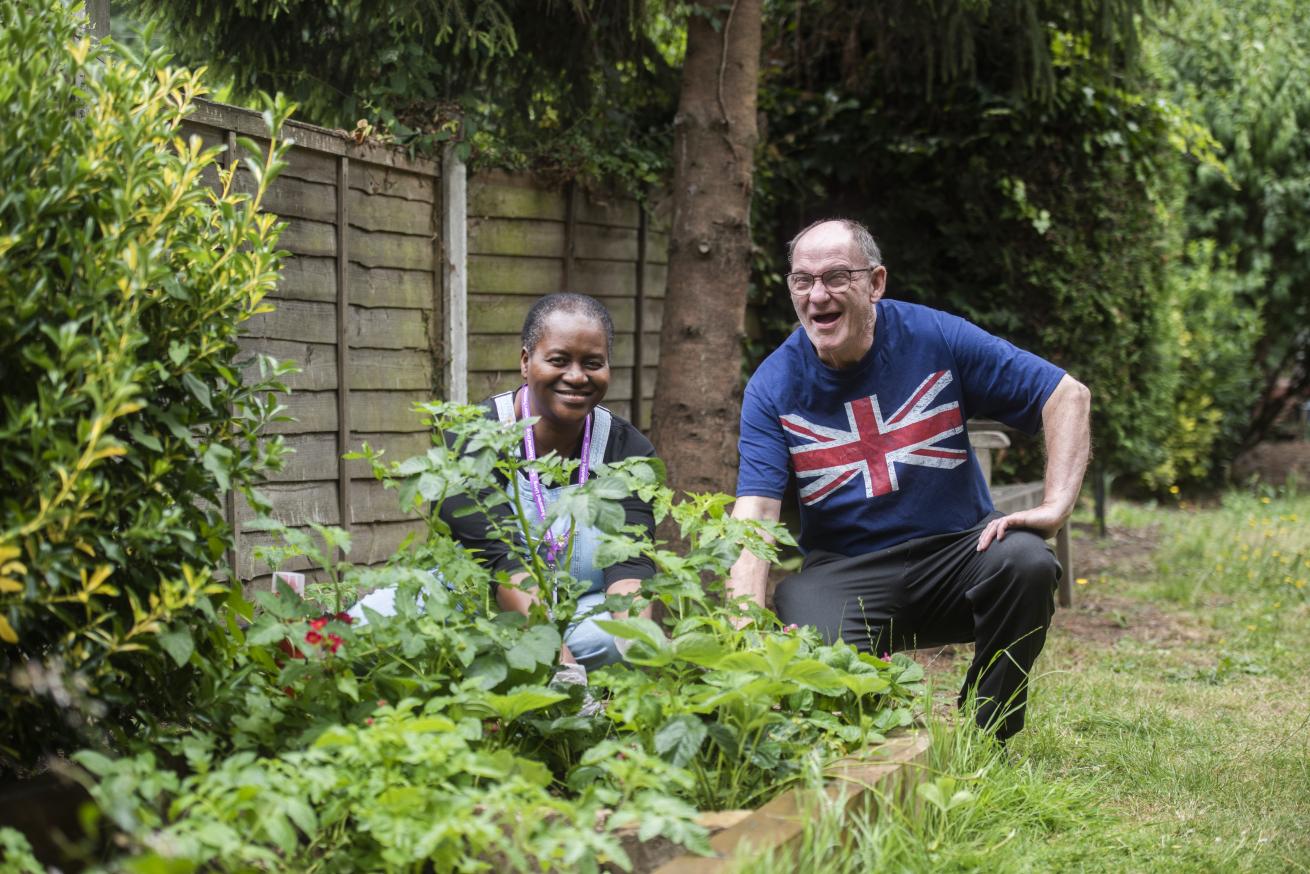 A woman and a man are kneeling beside a garden bed filled with various plants. The woman, wearing a purple apron, smiles as she tends to the garden. The man, wearing a blue shirt with a Union Jack design, is crouched down, looking at the plants with a cheerful expression. They are surrounded by greenery and a wooden fence in the background, creating a vibrant and lively outdoor setting.