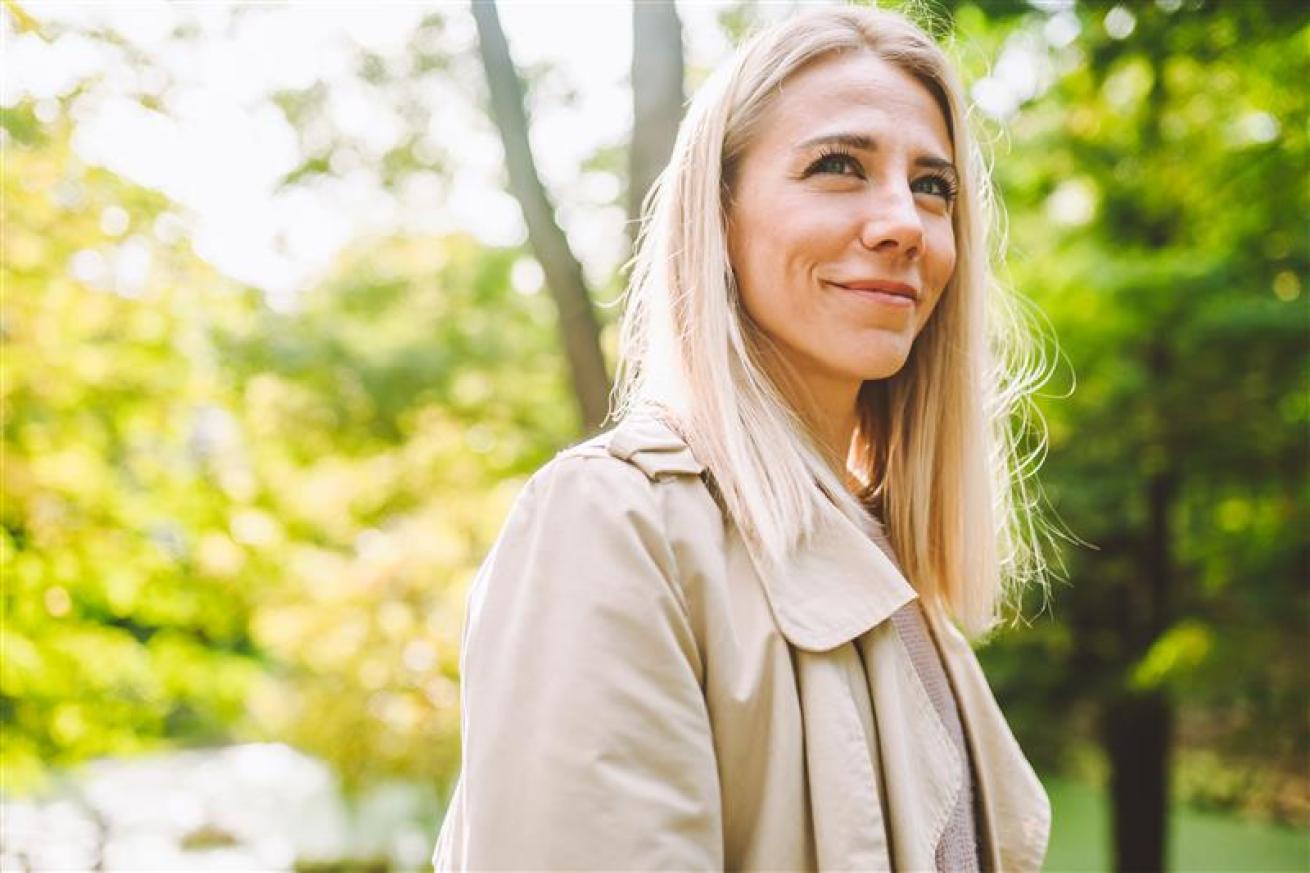 A representative photo of Lauren showing a person with long, straight blonde hair wearing a light beige trench coat, standing outdoors in a green, leafy park setting. Sunlight filters through the trees in the background, creating a bright and natural atmosphere.