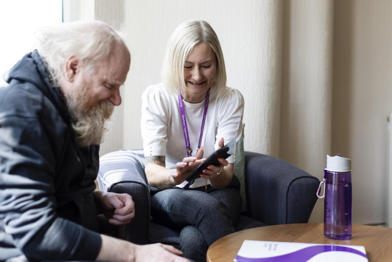 A woman with short blonde hair and a white shirt is sitting next to a man with a long beard, both smiling as they look at a smartphone together. They are seated on a couch in a well-lit room, with a water bottle and a document on a table in front of them. The atmosphere appears friendly and collaborative.