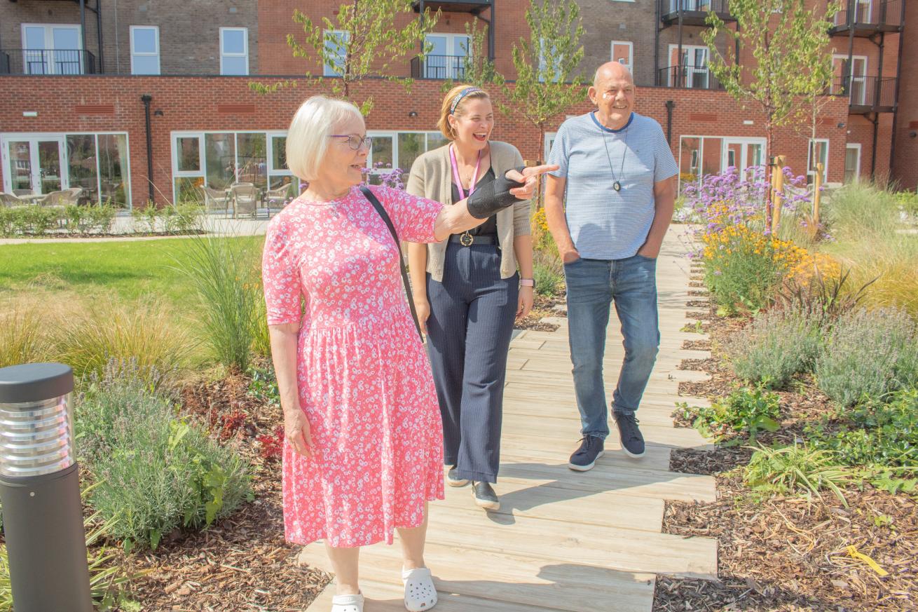 A group of three people is walking along a wooden pathway in a landscaped garden. The first person, a woman with short grey hair, is gesturing with her left hand. The second person, a woman in smart casual attire, is smiling and looking at the first woman. The third person, a man dressed in all blue, is walking beside them.
