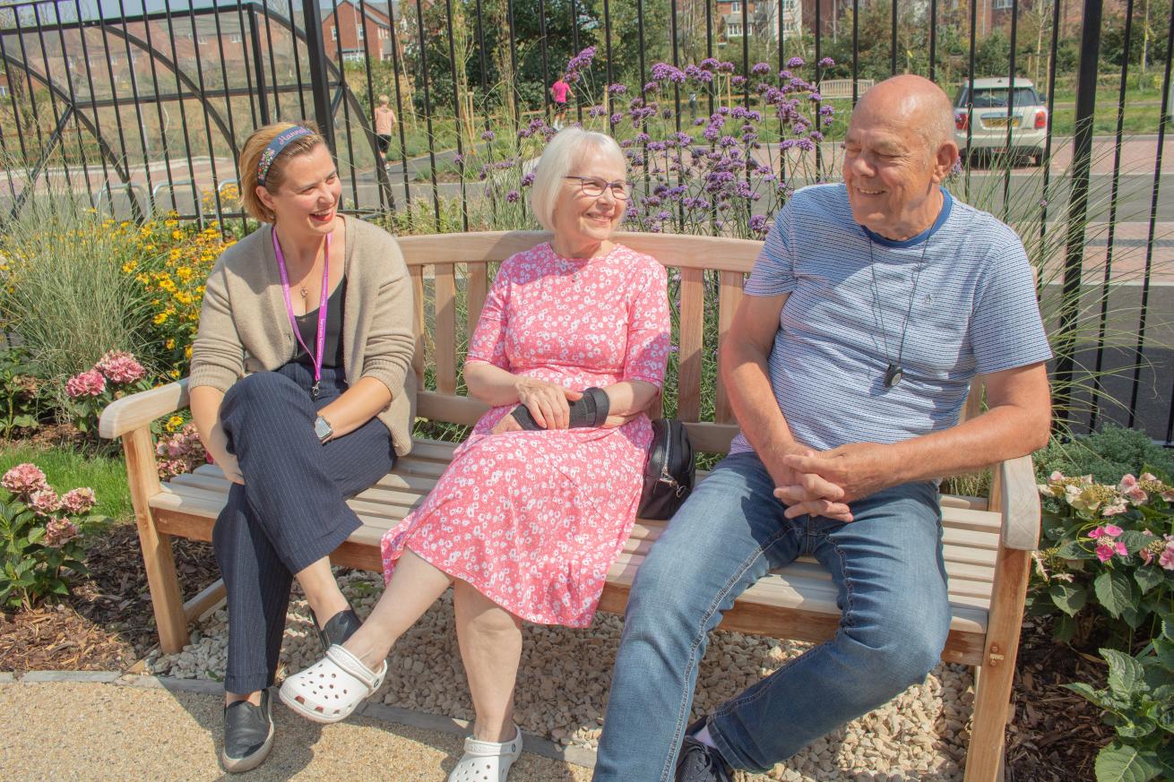 A staff member and two residents sit smiling on a bench in a floral garden.