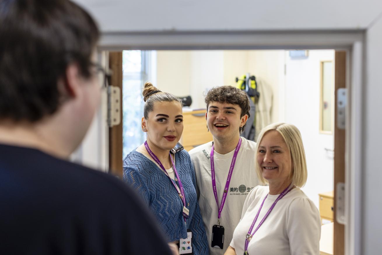 Three Sanctary Supported Living Staff members stand in a doorway smiling brightly. They are facing another person who is out of focus on the far left of the frame. 