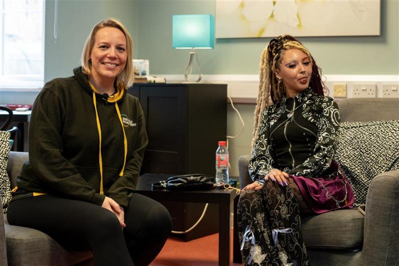 A woman with blonde hair wearing a black hoodie stands next to a woman with long, braided hair styled in a fashionable outfit. They are both smiling and posing for the photo in a well-lit room with a floral artwork on the wall. There are soft furnishings in the background, suggesting a cozy environment.