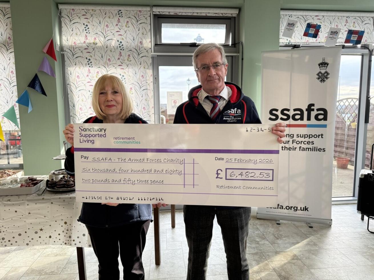 A man and a woman stand together in a bright, decorated room, holding a large novelty cheque made out to SSAFA - The Armed Forces Charity.