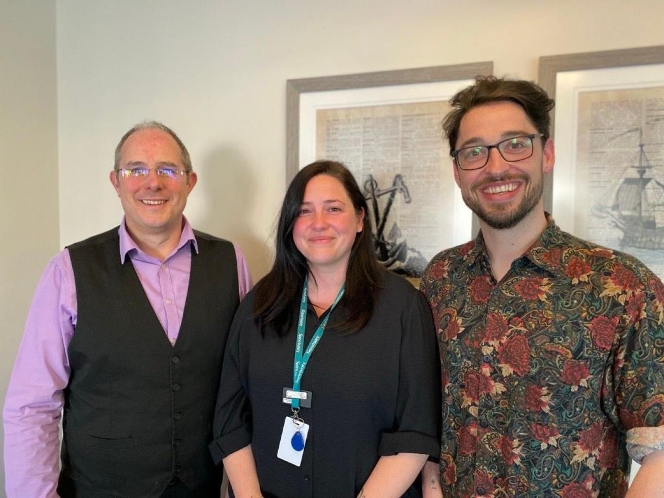 Three members of the Sanctuary Supported Living Learning and Development team standing indoors. One person wears a lilac shirt with a dark waistcoat, one wears a dark long‑sleeved top with a green lanyard, and one wears a patterned short‑sleeved shirt. They are standing side by side in front of framed artwork on a light‑coloured wall.