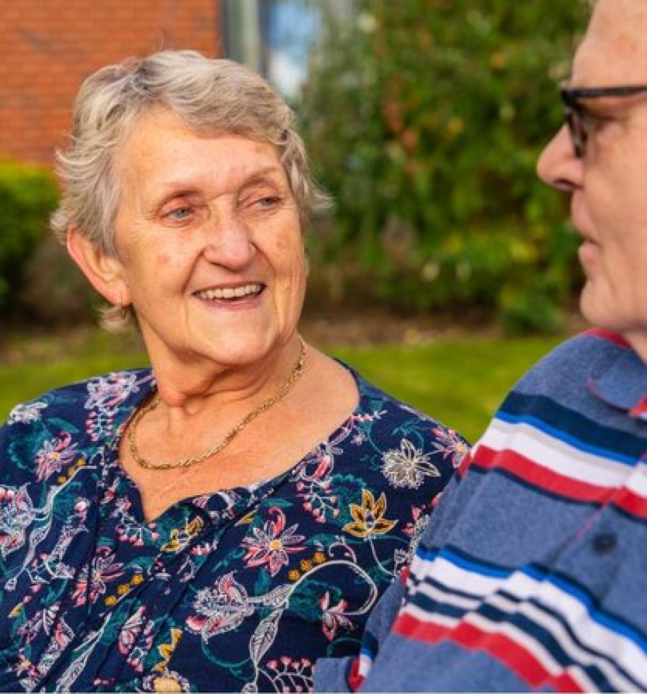 An elderly woman with short gray hair and a floral blouse smiles warmly at an elderly man wearing glasses and a striped shirt. They are sitting outdoors in a green, well-maintained garden, engaged in a friendly conversation. The background features shrubs and a brick building.