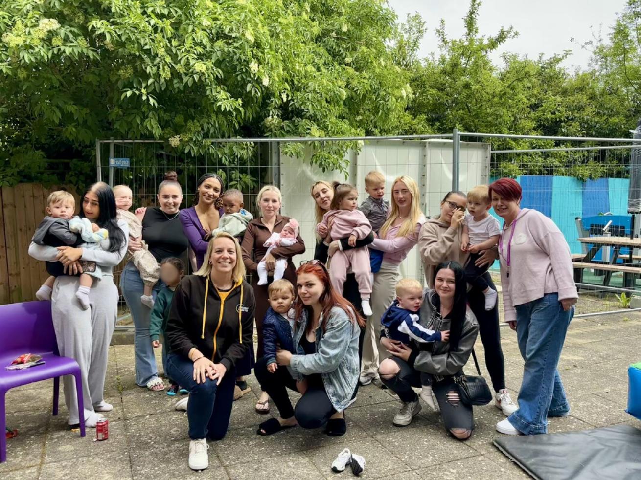 A group of women and children are gathered outdoors on a paved area. Some women are holding babies, while others are standing or kneeling with toddlers. The group is smiling and appears to be enjoying a casual gathering. In the background, there are trees, a fence, and some outdoor furniture. The atmosphere is relaxed and friendly.