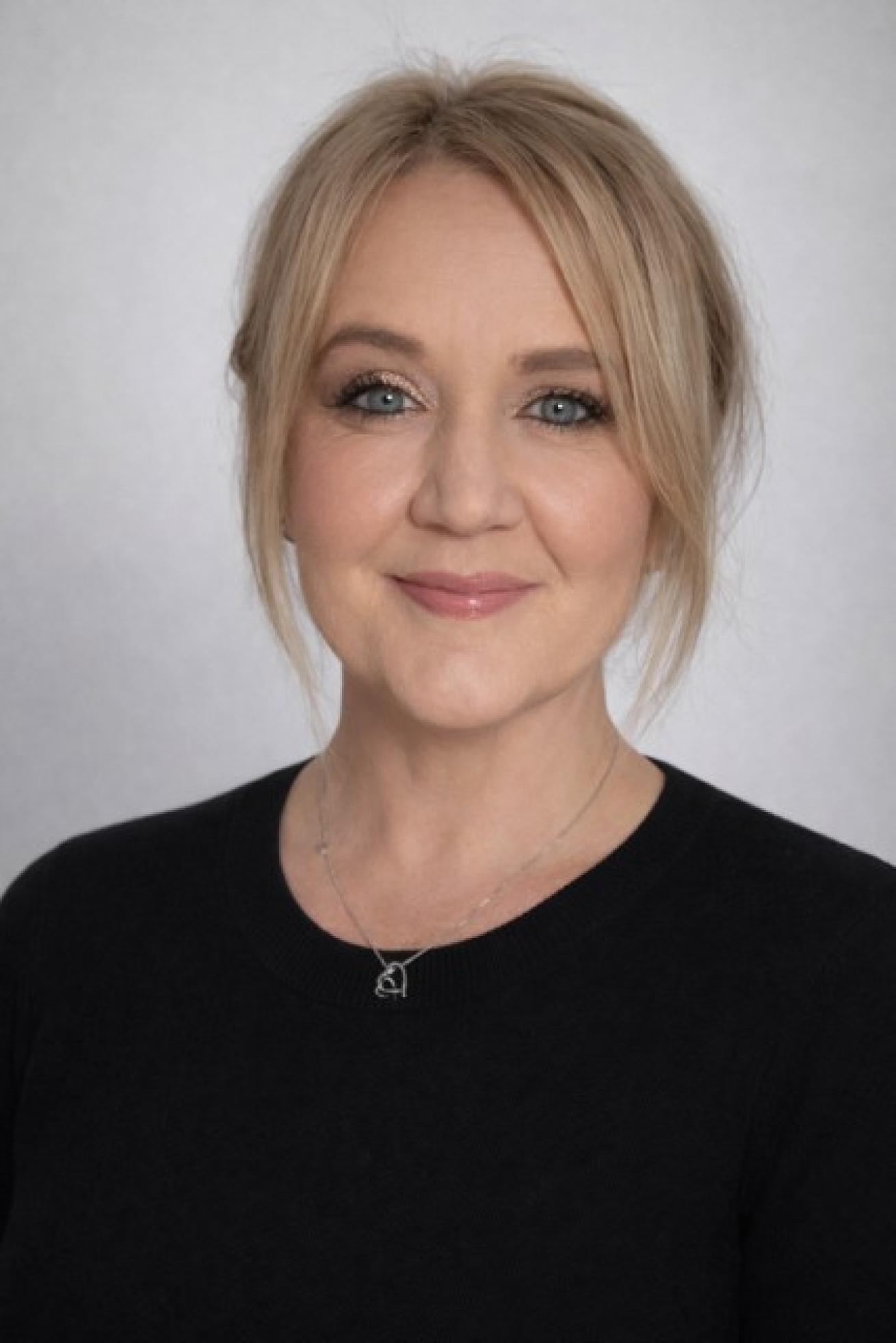A person with light blonde hair is smiling softly at the camera. They are wearing a black top and a delicate necklace. The hair is styled in a loose updo with some strands framing the face. The background is a plain, light gray color.