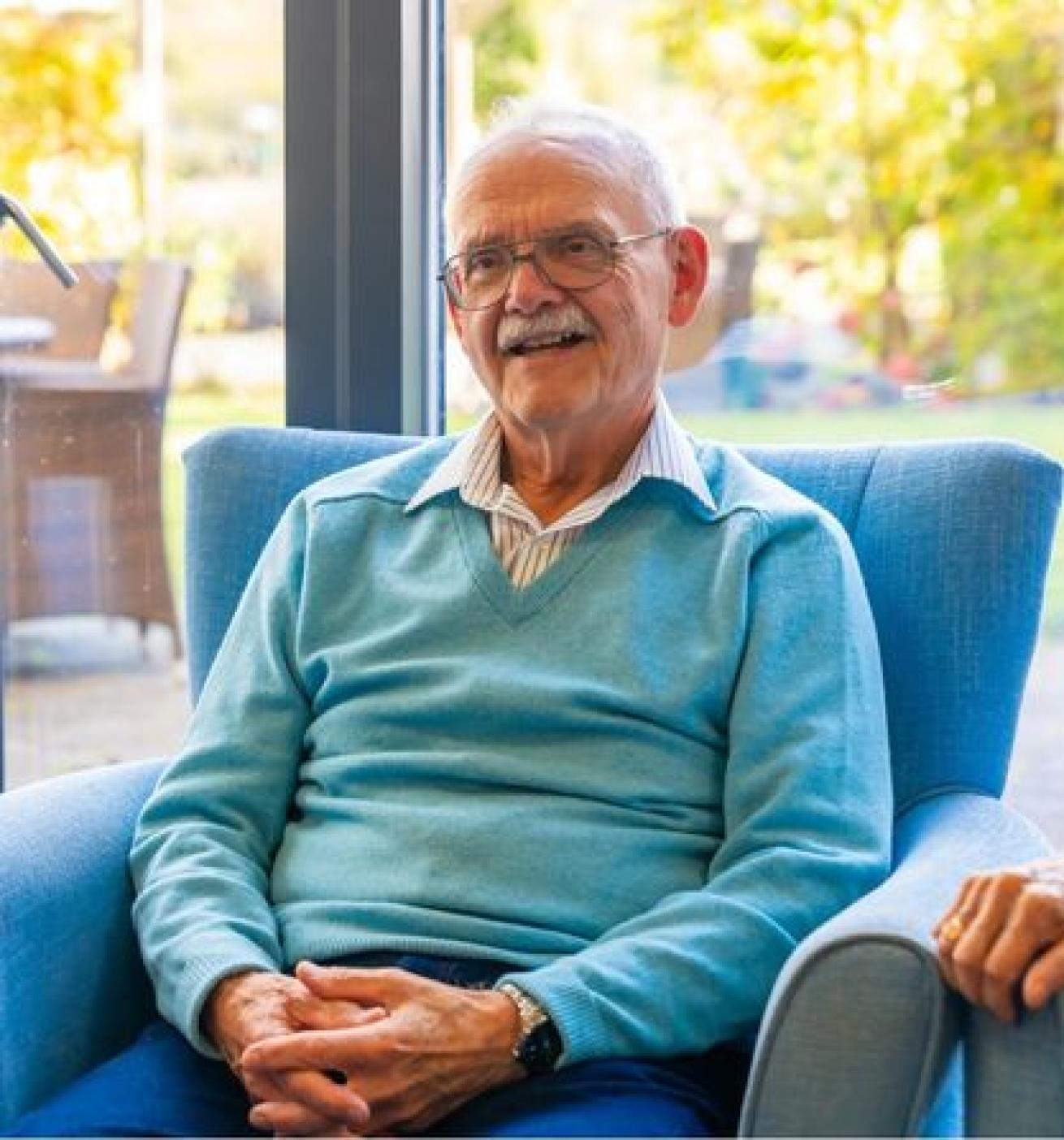 An elderly man with glasses and a light blue sweater sits comfortably in a blue armchair. He has a warm smile and is engaged in conversation. The background features a bright, sunny garden with greenery and outdoor furniture visible through large windows.