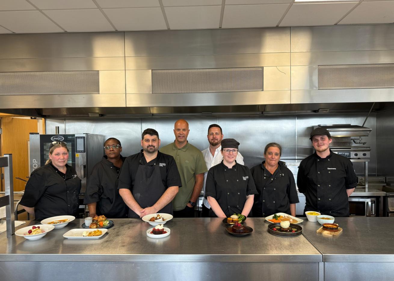 Catering colleagues stand behind a stainless-steel worktop displaying their completed dishes in a professional training kitchen.