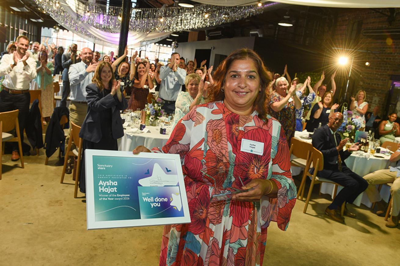 A woman in a red and white patterned dress stands smiling, holding a framed certificate. The certificate reads "Aysha Hajat, Employee of the Year." Behind her, a large group of people are standing and clapping in a decorated venue with draped fabric and lights. Tables are set with flowers and glasses, suggesting a celebratory event.