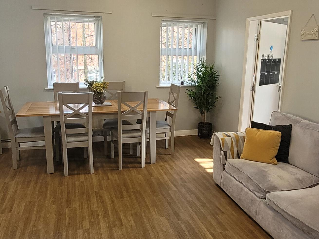 A bright and airy dining area featuring a wooden table surrounded by six light-colored chairs. A small potted plant is positioned near the table. To the right, a material grey sofa with a yellow and black cushion is placed against the wall. A wall-mounted organiser is visible in the background.