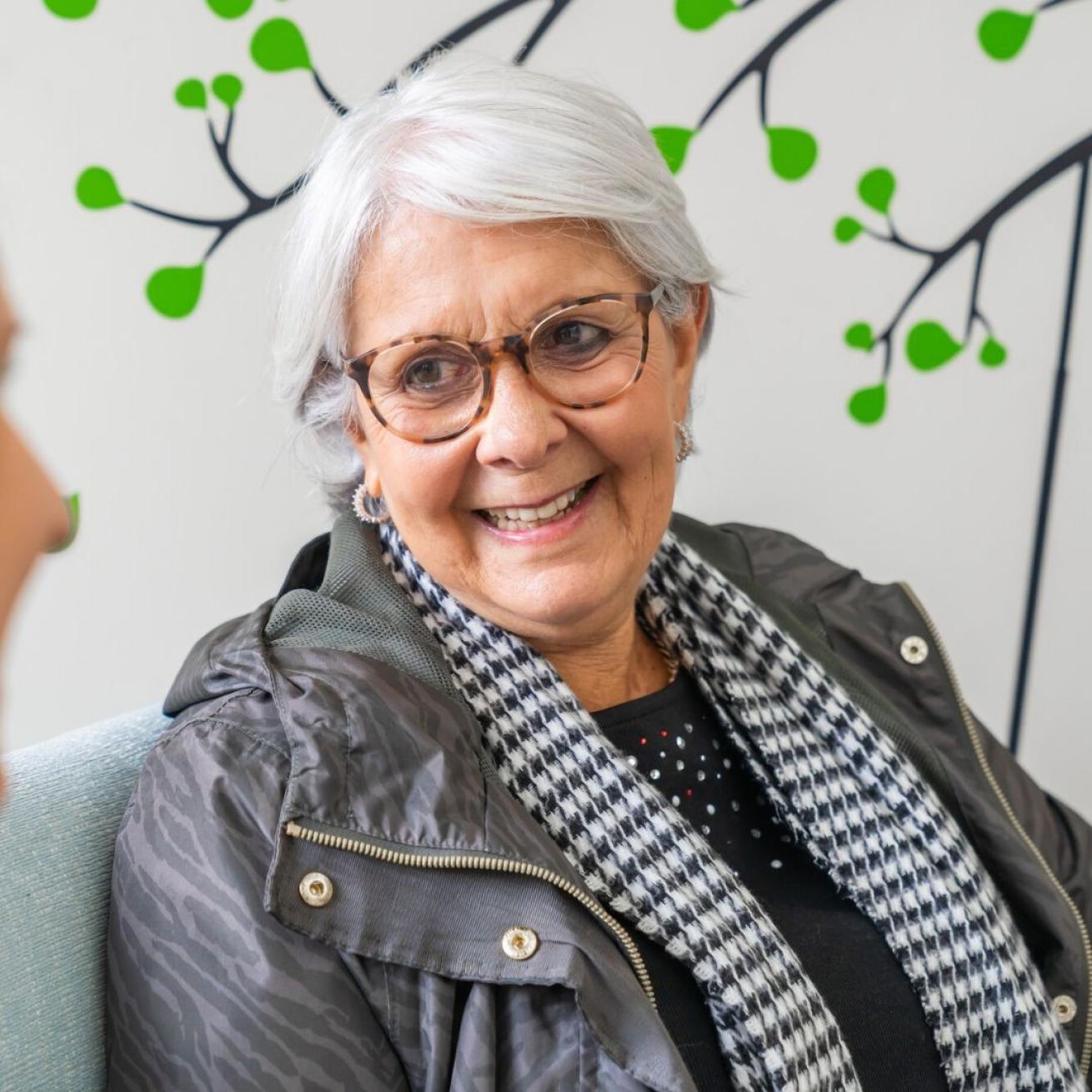 A smiling older woman with short gray hair and glasses is sitting in a cozy setting. She is wearing a dark jacket and a black and white checkered scarf. In the background, there is a wall decorated with green leaf motifs, creating a warm and inviting atmosphere. The woman appears to be engaged in conversation, looking attentively at the person she is speaking with.