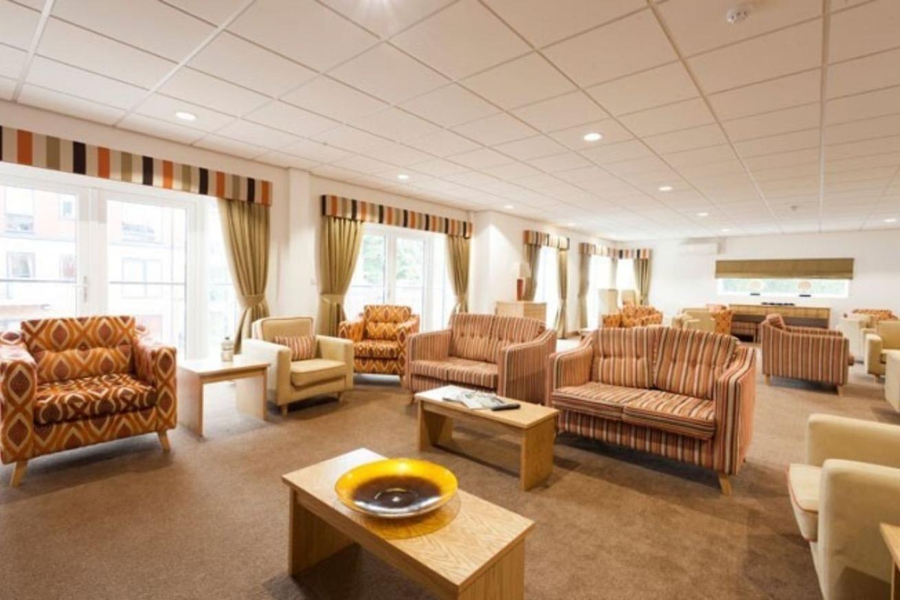 Interior view of a comfortable lounge area within a care facility. Several patterned armchairs and striped sofas are arranged around a wooden coffee table with a decorative bowl. Large windows provide natural light and a view of the outdoors. The room has a neutral color palette with a textured ceiling and patterned curtains.
