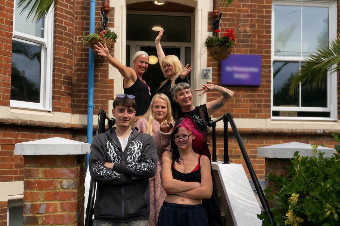 Group of six people standing on the steps outside a red brick residential building. Two people stand at the front with arms folded, while four others pose behind on the staircase. Hanging flower baskets frame the entrance with surrounding white-framed windows, black handrails and greenery.