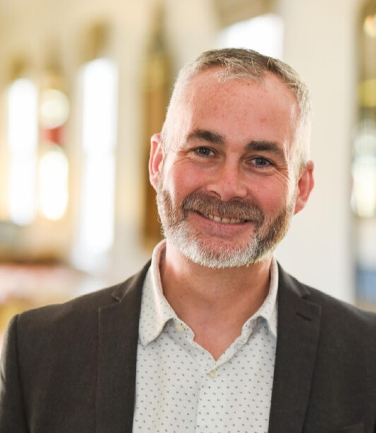 A smiling man with a beard is wearing a dark blazer over a light-colored, patterned shirt. He is standing in a well-lit indoor setting with soft, blurred background elements that suggest a warm and inviting atmosphere.