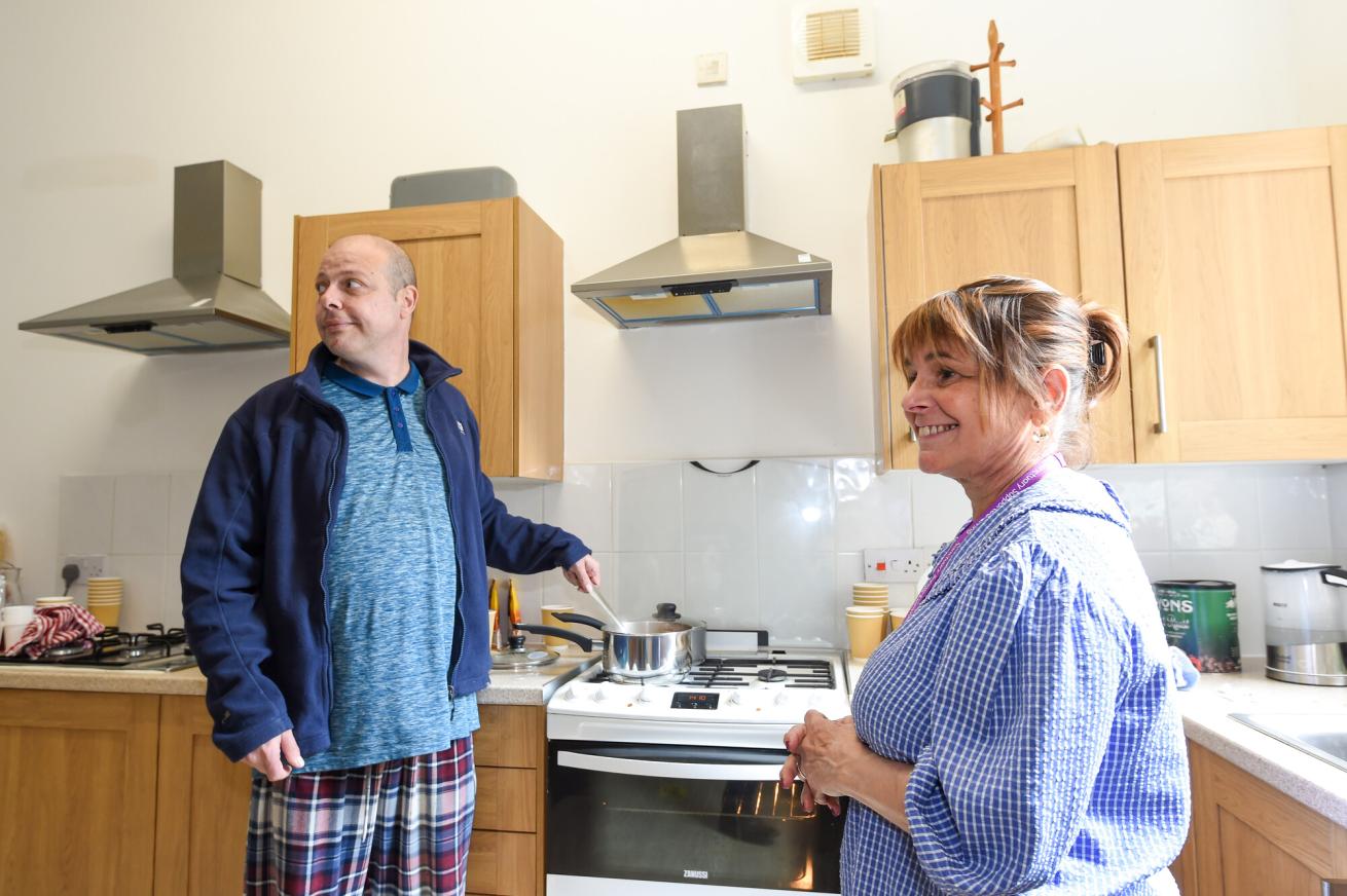 A man in a blue jacket and plaid pajama pants stands at a stove, stirring a pot. He looks to the side with a slight smile. A woman in a blue checkered shirt stands nearby, smiling back at him. The kitchen has wooden cabinets and modern appliances, with various kitchen items visible on the counter.