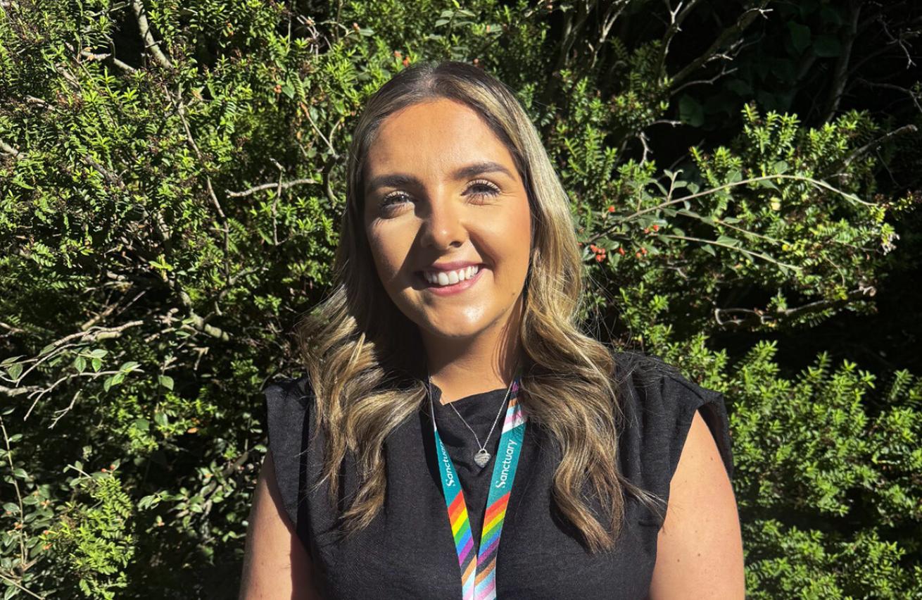 A young woman with long, wavy hair stands smiling in front of a lush green background. She is wearing a black top and a colorful lanyard with a rainbow design around her neck. The sunlight highlights her features, creating a warm and inviting atmosphere.