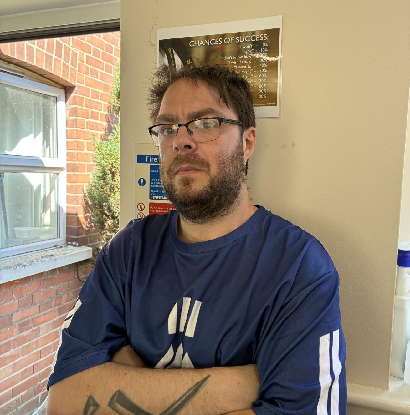 A man with glasses and a beard stands with his arms crossed, wearing a blue shirt with white stripes. A black geometric tattoo is visible on one forearm. He has a serious expression on his face. In the background, there is a poster on the wall that reads "CHANCES OF SUCCESS," and a window showing a glimpse of greenery outside.