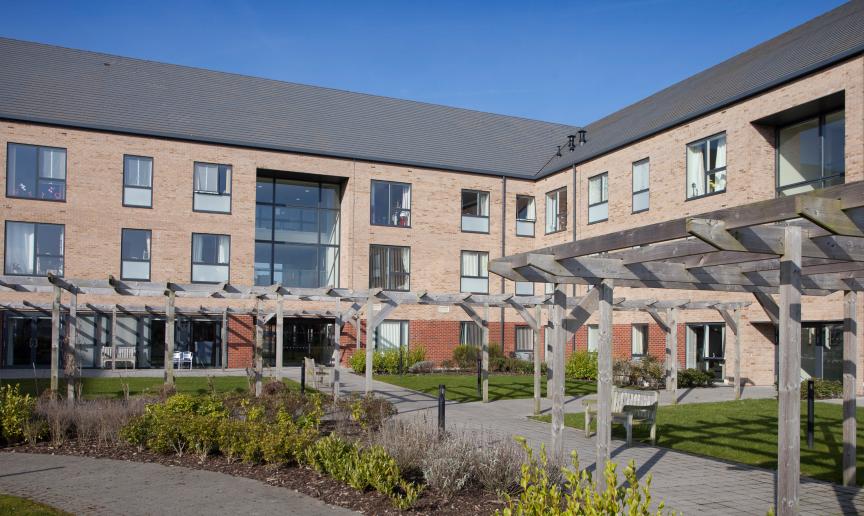 Stylized retirement living community with an array of large windows over looking a sectioned off grassed areas, flower beds and walkways, located in Stoke-on-Trent.