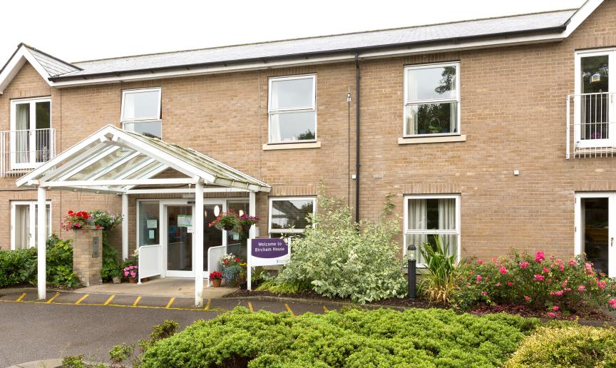 Two storey sandy brick-built retirement community located in Cambridgeshire. The entranceway has no step access. The building is decorated by a mixture of mature shrubbery and plants.