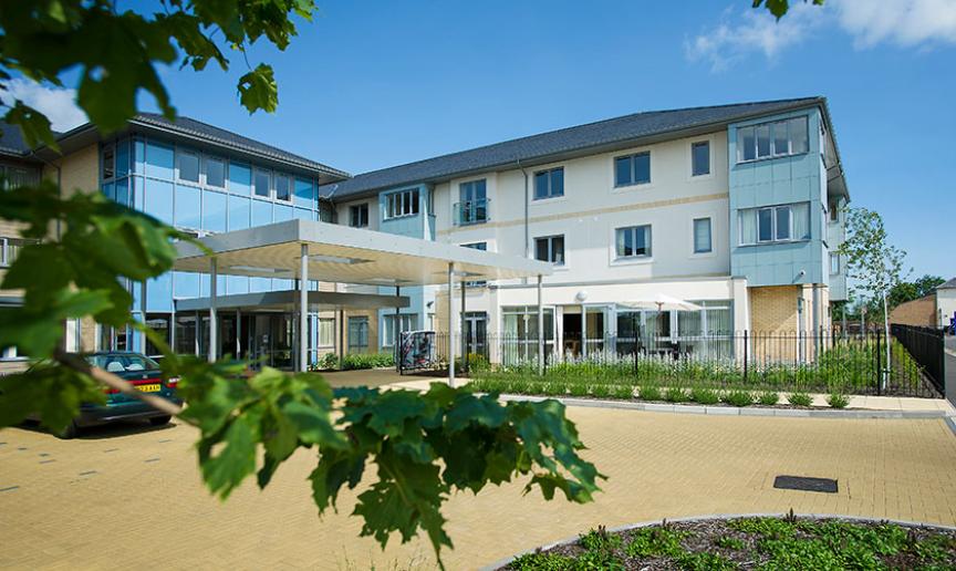 Contemporary, bright retirement living building. The bold blue pops of colour on a selection of the elevations gives a softer approach to the building against the white and grey. A small carpark features at the front of the property. 
