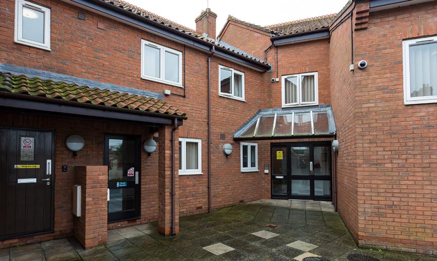 Multi-entranced brick exterior with terracotta clay roof tiles. Multiple security cameras and lighting. Accessed by a clean concrete tiled walkway to Drawbridge House.