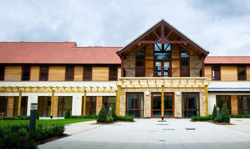 A luxurious property with a grand entrance and first storey balcony overlooking the white slabbed walkway. The property has been built using a mixture of natural materials such as brick, wooden panelling, and wooden beams. Small shrubbery plants line the perimeter.