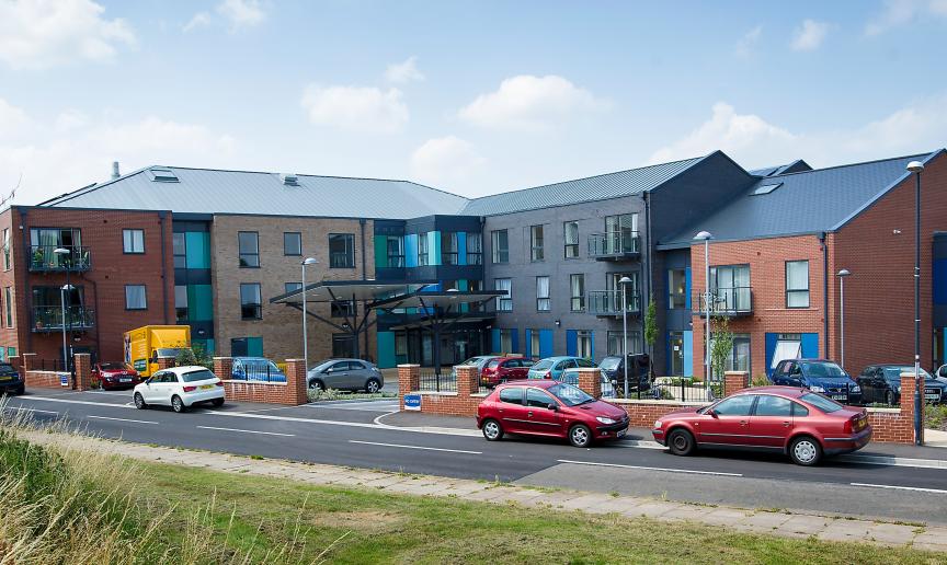 Located on a quiet corner plot the bold retirement living community façade consists of red, stone and grey brick and blue panelling on the larger exterior windows. Parking is located around the perimeter of the building.