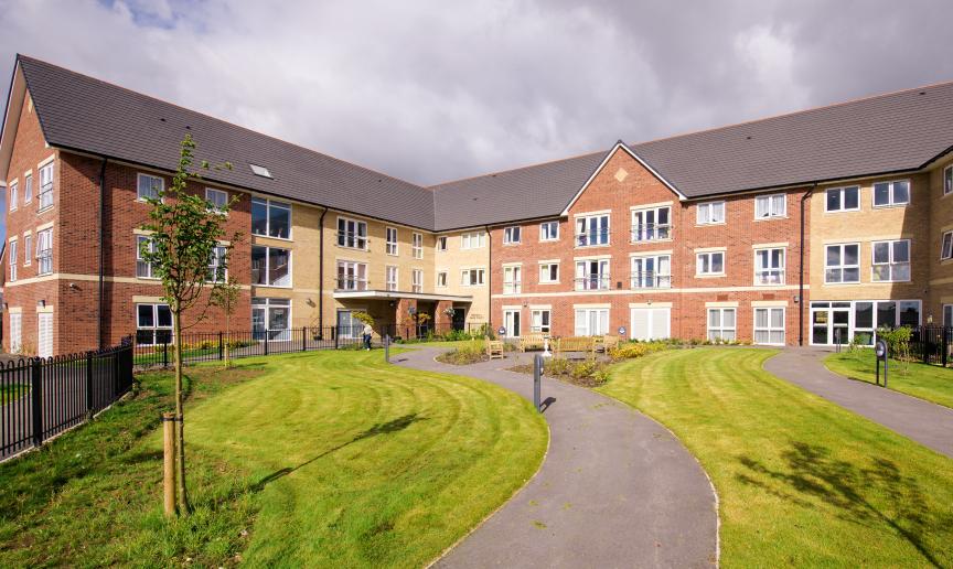 The newly built, yellow, and red bricked, three storey retirement living property set behind winding pathways which lead to a circular outdoor seating area as well the property and lovely grassed areas.