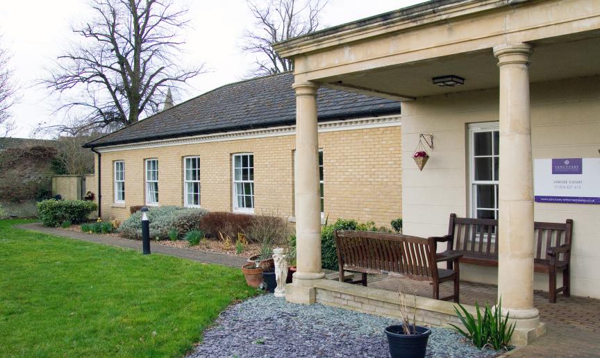 A period designed stone built building is home to the entrance of the property, the entrance is covered by a stone built porch with chunky columns supporting the roof. Two wooden benches sit underneath looking out onto the gardens. The rest of the building is built with soft coloured yellow bricks and long rectangular windows. 