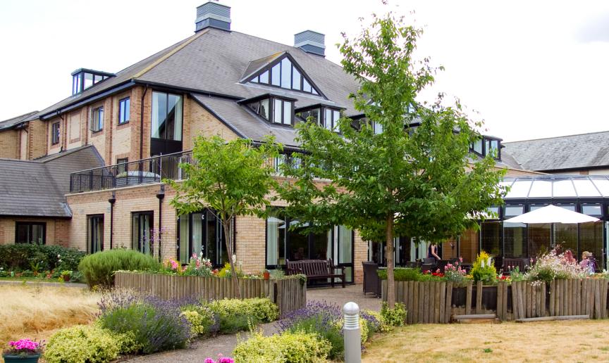 An idyllic brick built property with mass amounts of over sized windows and added skylights, set amongst a brick paved patio area and parasol seating enclosed in by wooden logged planters and colourful plants. 