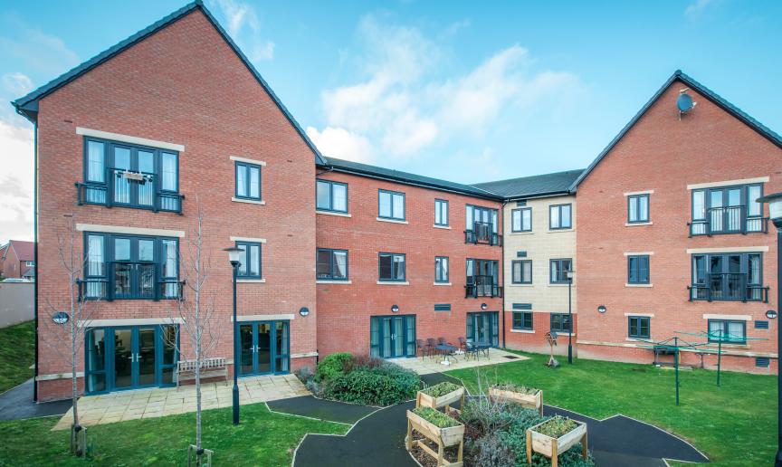A modern red bricked new build retirement property. The building is built as a three storey corner plot with a small patio area along the left hand side of the property hosts an array of seating options from a wooden bench or metal chairs and circular tables. The patio leads into a circular tarmacked area with wooden planters and shrubbery, the rest of the garden is covered in grass.  