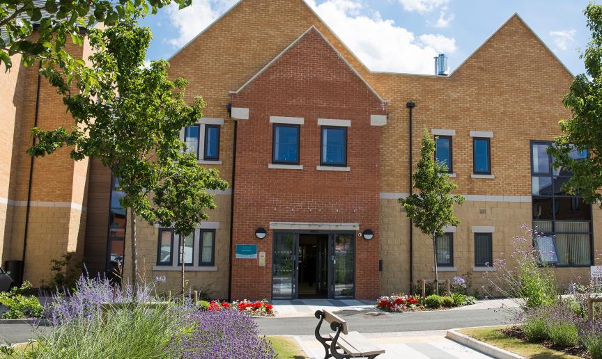 New build yellow brick built two storey property, with exaggerated triangular roofing, the entranceway is protruding the exterior designed in a red brick with mimicking pointed triangular roof. The outdoor area is planted with planted trees, flowers, and mature shrubbery.
