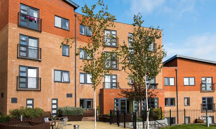 Beautiful contemporary apartment building contrasting dark grey window frames with the multitoned red bricked exterior. The outdoor area offers metal seating as well as numerous wooden benches all looking out to the newly planted trees and garden area.