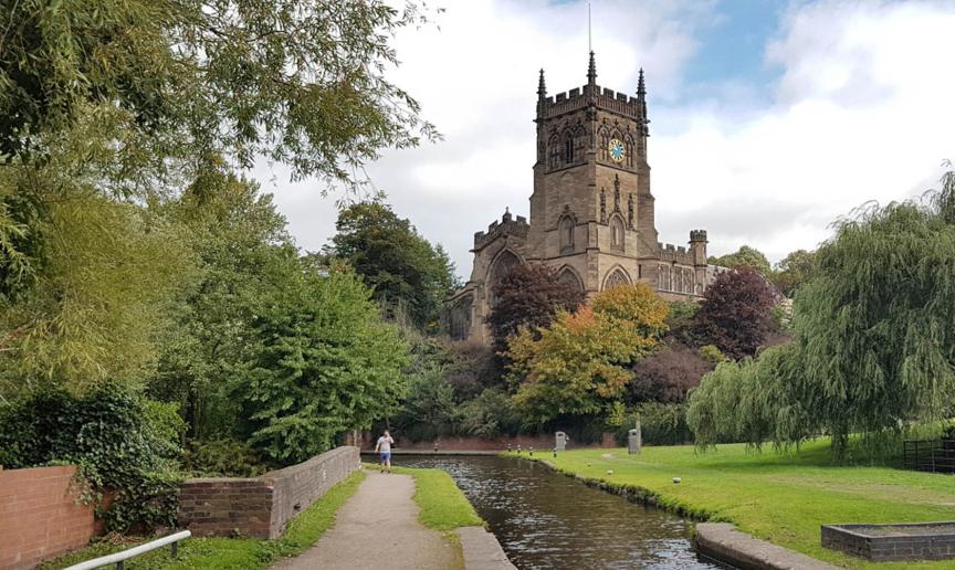 Canal footpath with grass verge and an array of mature trees with a 15th century church towering above.